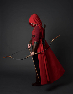 穿着红色中世纪服装和斗篷的黑发女孩的全长肖像，手持弓箭。 站在灰色工作室背景上的姿势。