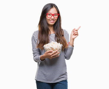 年轻的亚洲女人在孤立的背景下吃爆米花，非常高兴地用手和手指指向一边
