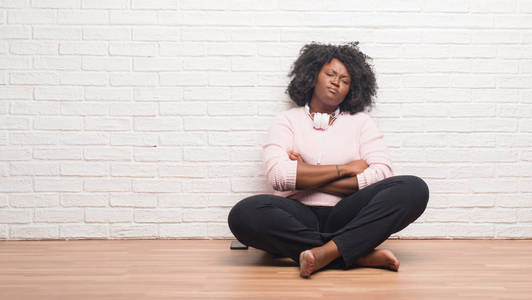 年轻的非裔美国妇女坐在地板上戴着耳机，怀疑和紧张的表情，脸上交叉的手臂。 消极的人。