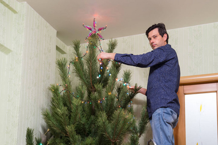 英俊的中年男子装饰了一棵圣诞树，一颗星星和一个五颜六色的灯泡花环站在客厅内部的梯子上。 新年假期概念舒适的家