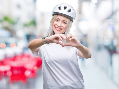 年轻的金发女人戴着自行车安全头盔，在孤立的背景上微笑，在爱中，用手显示心脏的象征和形状。 浪漫的概念。