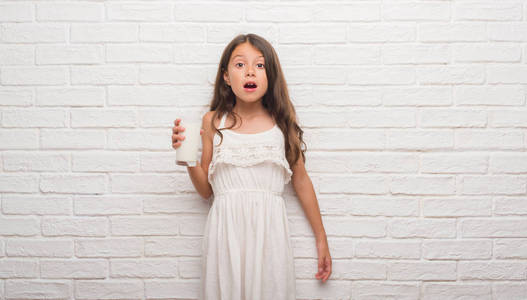 的西班牙裔小孩在白砖墙上喝着一杯牛奶,吓了一跳,脸上带着惊讶的表情