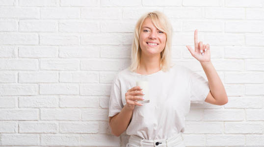 成年白种人妇女在白色砖墙上喝着一杯牛奶，惊讶地想到了一个主意或问题，用手指指着幸福的脸第一