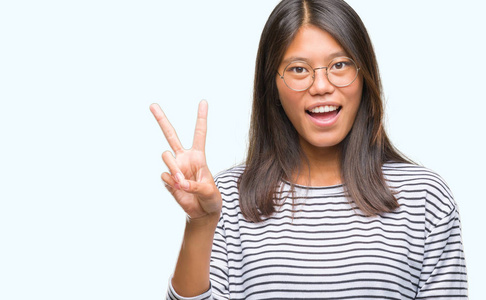 年轻的亚洲女人戴着眼镜，在孤立的背景上微笑，快乐的脸对着镜头眨眼，做胜利的标志。 二号。