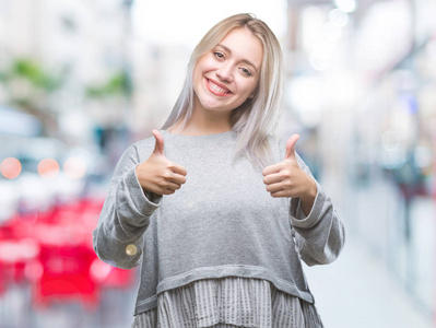 年轻的金发女人在孤立的背景下，赞成做积极的手势，用手竖起大拇指，微笑，并为成功而高兴。 看着相机赢家的手势。