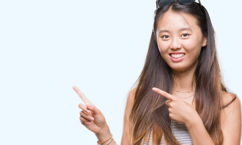 年轻的亚洲女人戴着太阳镜在孤立的背景上微笑着，看着相机，用两只手和手指指向一边。