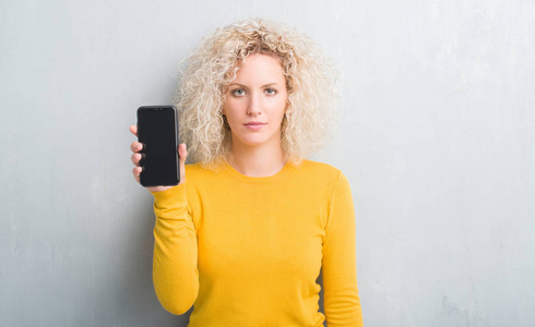年轻的金发女人，在灰色的灰色背景下，显示着空白的智能手机屏幕，智能面孔上充满自信的表情，严肃的思考