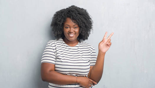 年轻的非裔美国人加大小的女人在灰色的格朗格墙上微笑，快乐的脸向相机眨眼，做胜利标志。 二号。
