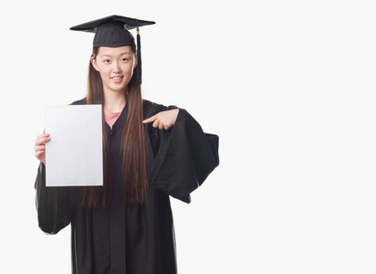 年轻的中国妇女穿着毕业制服，拿着纸学位，惊讶地指着自己