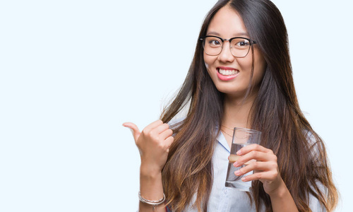 年轻的亚洲女人喝着一杯水，在孤立的背景上指着，用拇指向旁边露出快乐的脸，微笑着