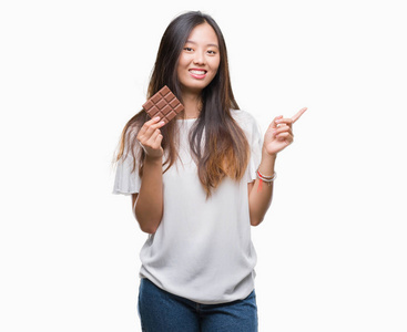 年轻的亚洲女人在孤立的背景上吃巧克力棒，非常高兴地用手和手指指向一边