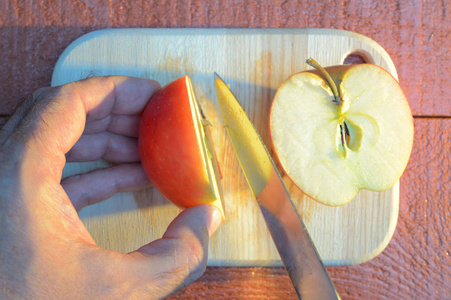 在日落时分,用一只手在一块红棕色背景的木切割板上用刀切苹果照片