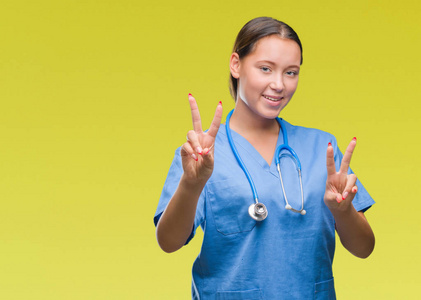 年轻的高加索医生女人穿着医疗制服，在孤立的背景上微笑着，看着镜头，手指在做胜利的标志。 二号。