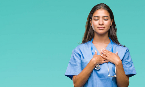 年轻的阿拉伯医生外科医生女士在孤立的背景上微笑，双手放在胸部，闭着眼睛，脸上有感激的手势。 健康概念。