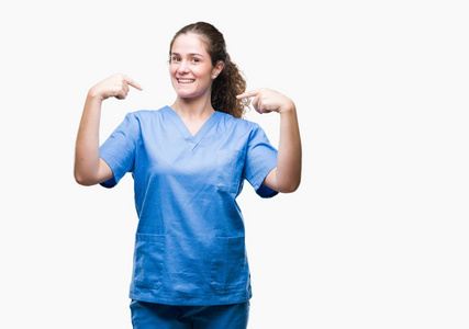 年轻的黑发医生女孩穿着护士或外科医生制服，在孤立的背景上看着自信，脸上带着微笑，手指着自己，骄傲和快乐。