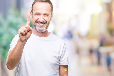中年老人穿着白色T恤，在孤立的背景上显示和指向第一手指，同时微笑自信和快乐。