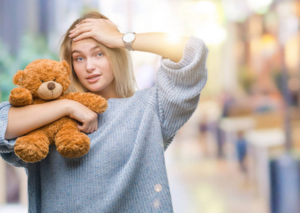 年轻的高加索女人抱着可爱的泰迪熊，在孤立的背景下，用手压着头，羞愧和惊讶的脸，愤怒和沮丧。 因为错误而恐惧和不安。