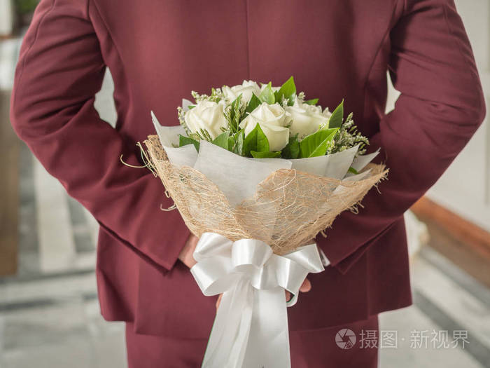 新郎穿着红色或勃艮第婚礼服背后藏着一束白色美丽的玫瑰花