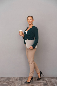 一个满脸笑容的年轻女商人，身披灰色背景，手提电脑，走路，喝咖啡