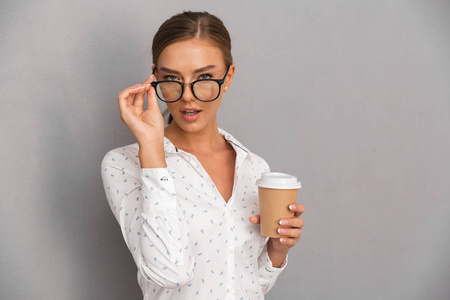 美丽的商务女性站在灰色的墙壁背景上，戴着眼镜喝着咖啡。