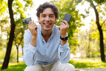 兴奋的年轻休闲男子手持手机，坐在城市公园里，出示信用卡