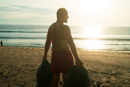 男子游客站在海滩上的后景，带着收集的垃圾袋在海上和日落的背景下