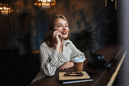 年轻开朗的女人，穿着条纹风衣，在手机上聊天，愉快地看着相机，带着小黑手提包和咖啡，在现代咖啡馆里消磨时间