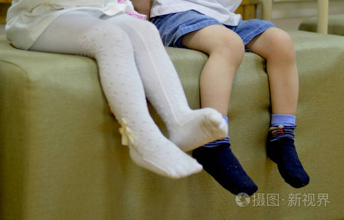 穿着白色连裤袜的女孩和坐在搁脚凳旁边的穿短裤和袜子的男孩的腿