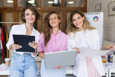 一群年轻漂亮的时尚女性，带着笔记本电脑和文件夹，快乐地看着镜头，一起在现代舒适的办公室工作。