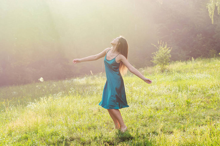美丽的微笑的年轻女子正在跳舞，在日落时赤脚在花草地上旋转。 日落时分，一位年轻的美女在森林附近的一片花草地上举起双手享受阳光