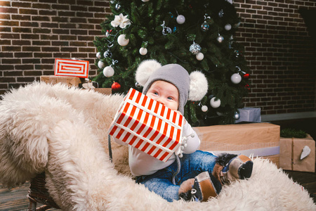 主题儿童圣诞节新年。 白种人小有趣的婴儿男孩，1岁，坐雪橇，熊皮，圣诞树头，温暖的帽子，巴拉邦球，球和牛仔裤周围的盒子礼物在晚上