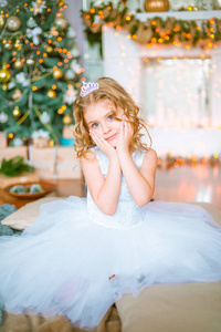一个可爱的小女孩，家里有一头金发，靠近一棵圣诞树，里面有礼物和花环，还有一个壁炉，坐在格子和枕头上