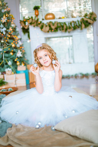 一个可爱的小女孩，家里有一头金发，靠近一棵圣诞树，里面有礼物和花环，还有一个壁炉，坐在格子和枕头上