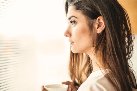 年轻的黑发女人拿着白色咖啡杯站在窗前