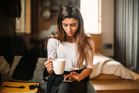 女人拿着白色咖啡杯，坐在室内使用智能手机