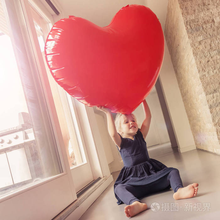 着蓝色裙子的金发小女孩坐在靠近窗户的地板上抱着一颗红色的充气心脏