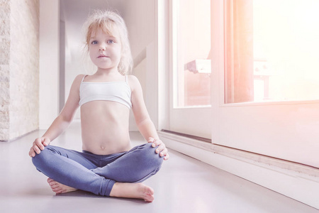 穿着白色上衣和蓝色紧身裤的金发小女孩在阳光明媚的房间里做瑜伽运动