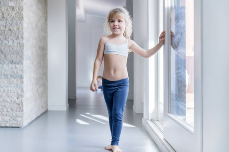 穿着白色上衣和蓝色紧身裤的金发小女孩站在靠近窗户的房间里照片