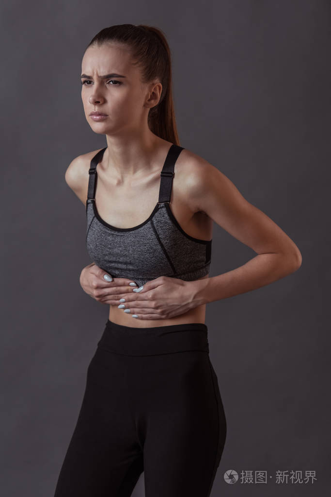 穿着运动服的漂亮女孩摸着她的肚子感到疼痛灰色背景