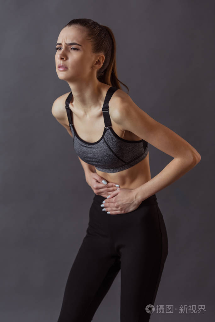 穿着运动服的漂亮女孩摸着她的肚子感到疼痛灰色背景