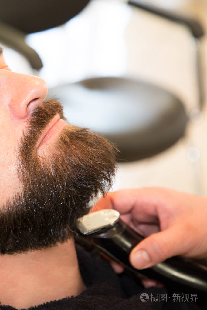 理发店里坐着的一个留着胡子的年轻人,由专业理发师修剪胡须