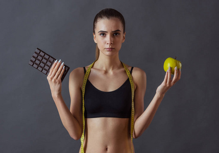 穿着运动服的漂亮女孩拿着苹果和巧克力棒，看着灰色背景的照相机