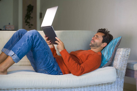 年轻,快乐,有魅力的男人,在现代公寓客厅里轻松地用笔记本电脑工作,躺