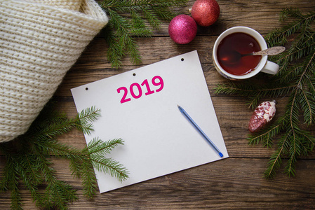 美丽的圣诞图片布置明信片与2019年新年一杯茶，一个笔记本，一支笔，计划，冷杉树枝，玩具，温暖的围巾，木制的乡村背景，俯视