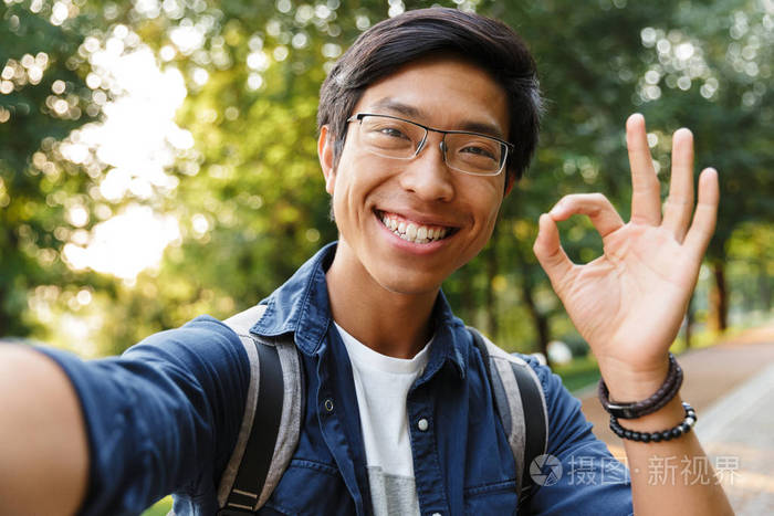 微笑的亚洲男学生戴着眼镜自拍在户外看相机时显示ok标志