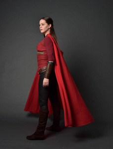 穿着红色中世纪服装和斗篷的黑发女孩的全长肖像。 站在灰色工作室背景上的姿势。