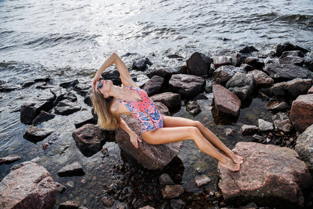美丽的金发女郎戴着太阳镜和泳衣站在岩石海滩上，白天
