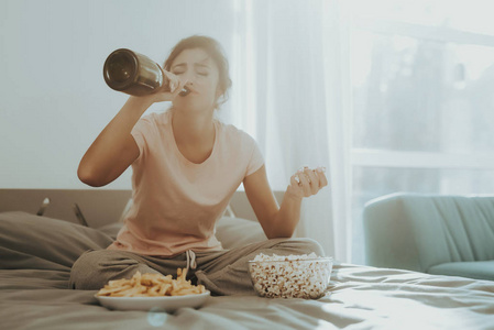 孤独哭泣的女人在床上喝香槟。 不愉快的假期。 悲伤的周末。 眼泪在脸上。 晴天。 独自在卧室里。 失望的女孩。 炸薯条和爆米花。