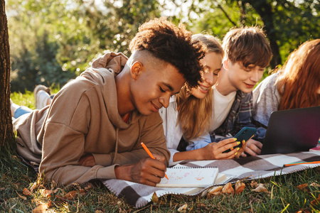 一群微笑的骡子学生在公园里一起做作业，他们用笔记本电脑和手机躺在草地上