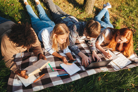 一群快乐的族学生在公园里一起做作业，用笔记本电脑躺在草地上